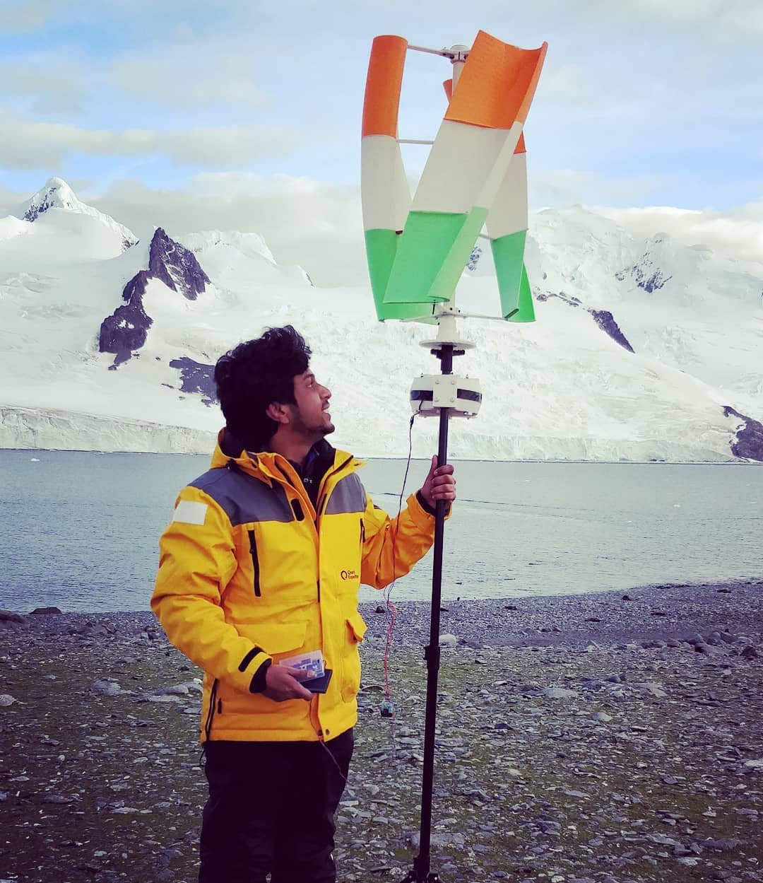 Vijay Raghav Varada in a yellow expedition parka on an Antarctic shore, holding a mast topped by a 3D printed vertical axis wind turbine painted in the Indian tricolour, with a glacier behind.