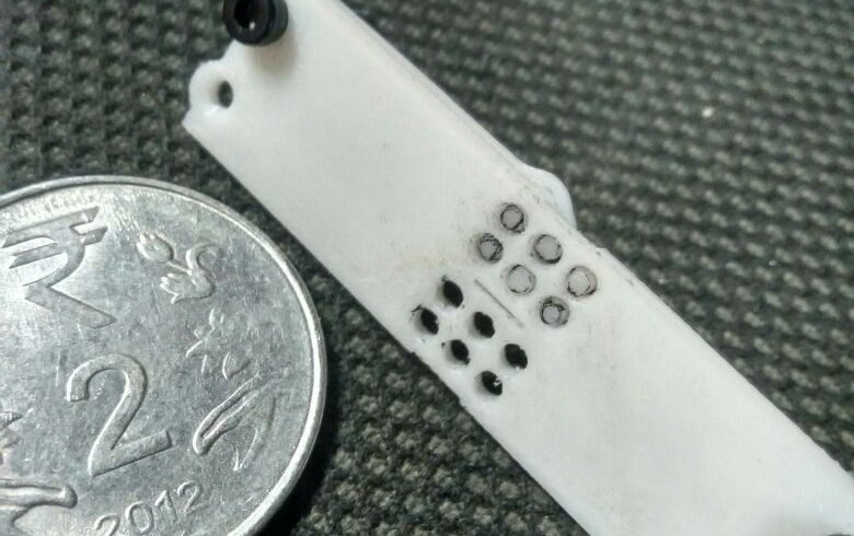 A printed Braille cell lying beside a two rupee coin, showing that the whole cell is barely longer than the coin is wide.