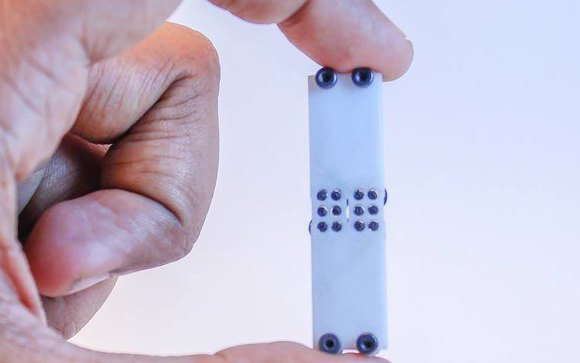 A white 3D printed Braille cell held between finger and thumb, its raised dots visible against the plastic.