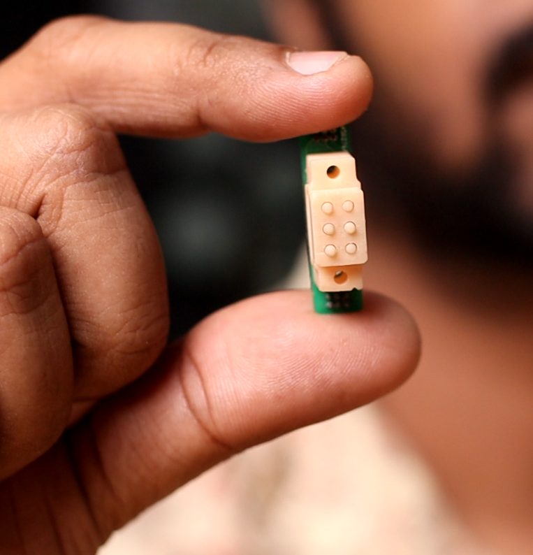 An assembled Braille module on its circuit board, held between finger and thumb.