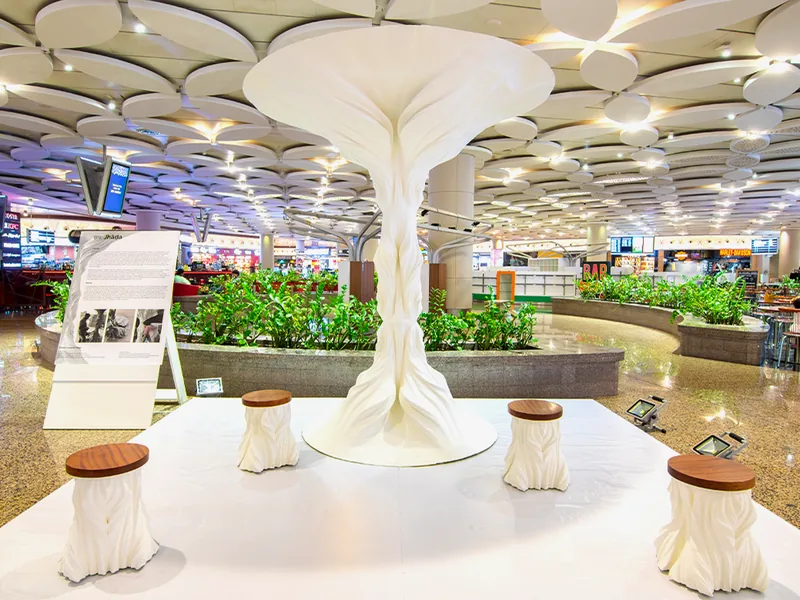 Jhada, a white 3D printed sculpture shaped like a tree, installed under the ceiling of Mumbai airport's Terminal 2 with printed stools around it.
