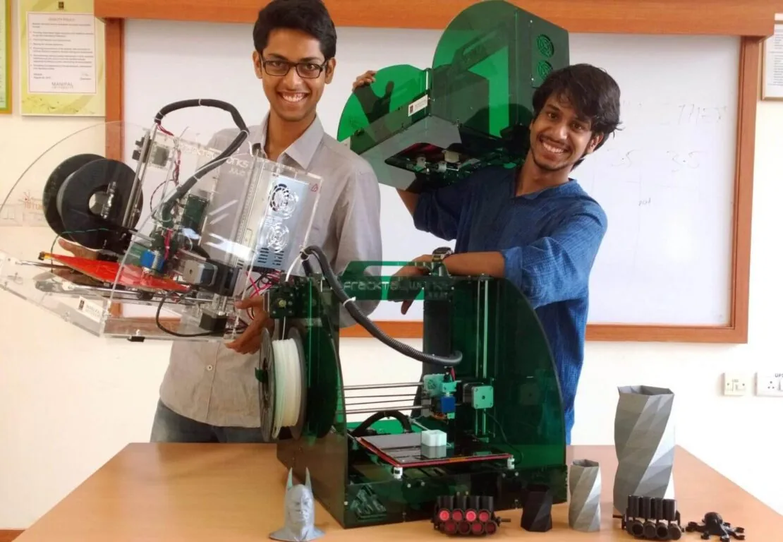Rohit Asil and Vijay Raghav Varada as students at Manipal, each holding an early Julia 3D printer built from clear and green acrylic, with printed test parts on the table in front of them.