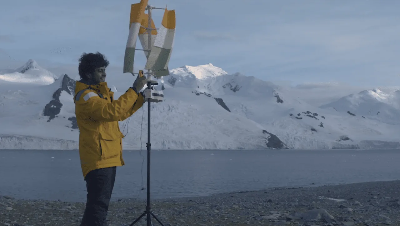 Vijay in a yellow parka adjusting the tricolour printed turbine and its generator on a mast, on the shore at Portal Point with Antarctic glaciers behind.