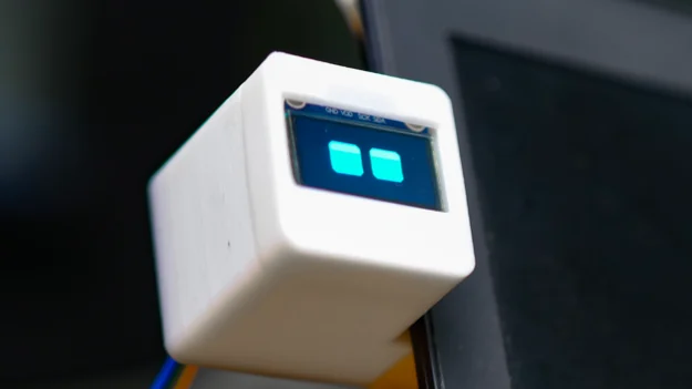 DeskBot, a small white 3D printed box with a glowing two-eyed screen, clipped to the edge of a monitor.