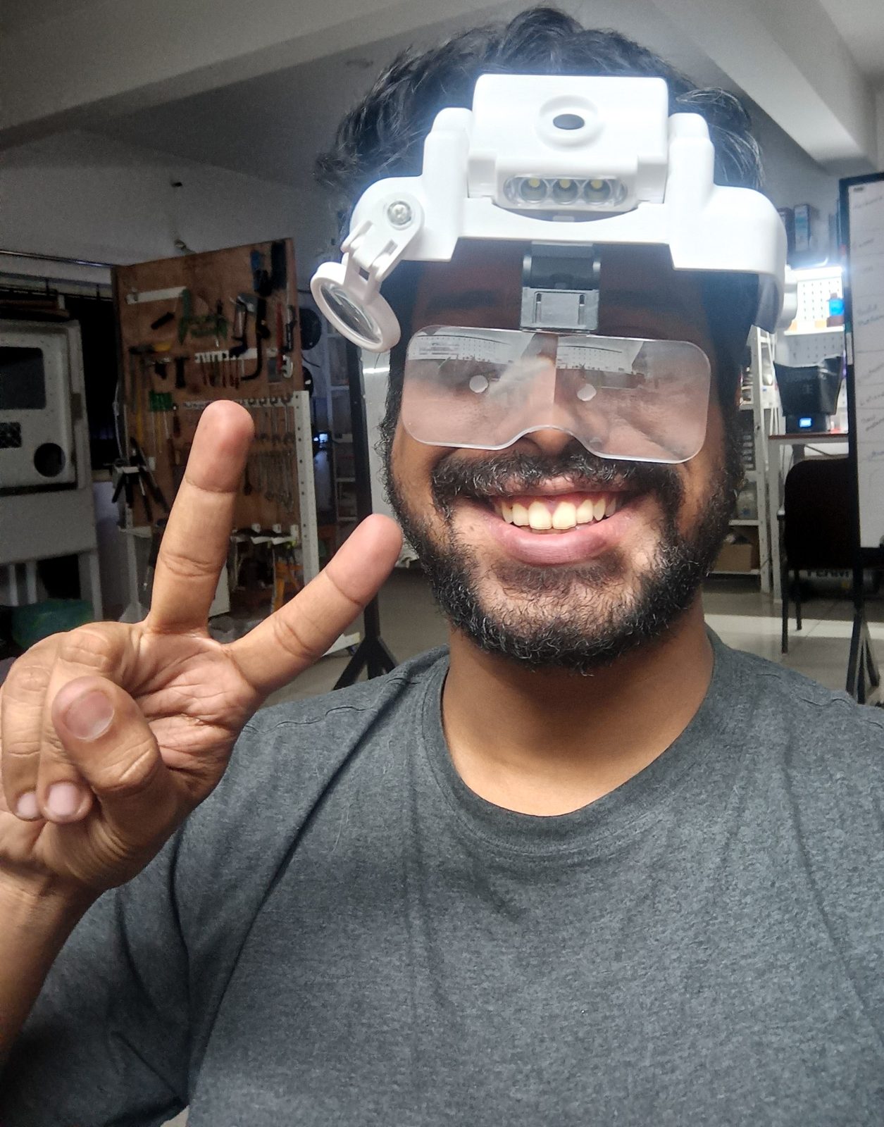 Vijay Raghav Varada in his workshop wearing a magnifier headset, with a tool wall and 3D printers behind him.