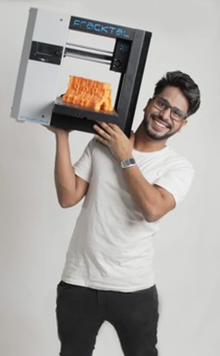 Vijay Raghav Varada carrying a Fracktal 3D printer on one shoulder, an orange print still on its bed.