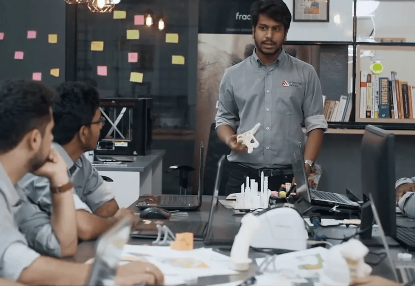Vijay Raghav Varada standing at the head of a workshop table, holding up a printed part while engineers listen, a Fracktal printer on the bench behind them.