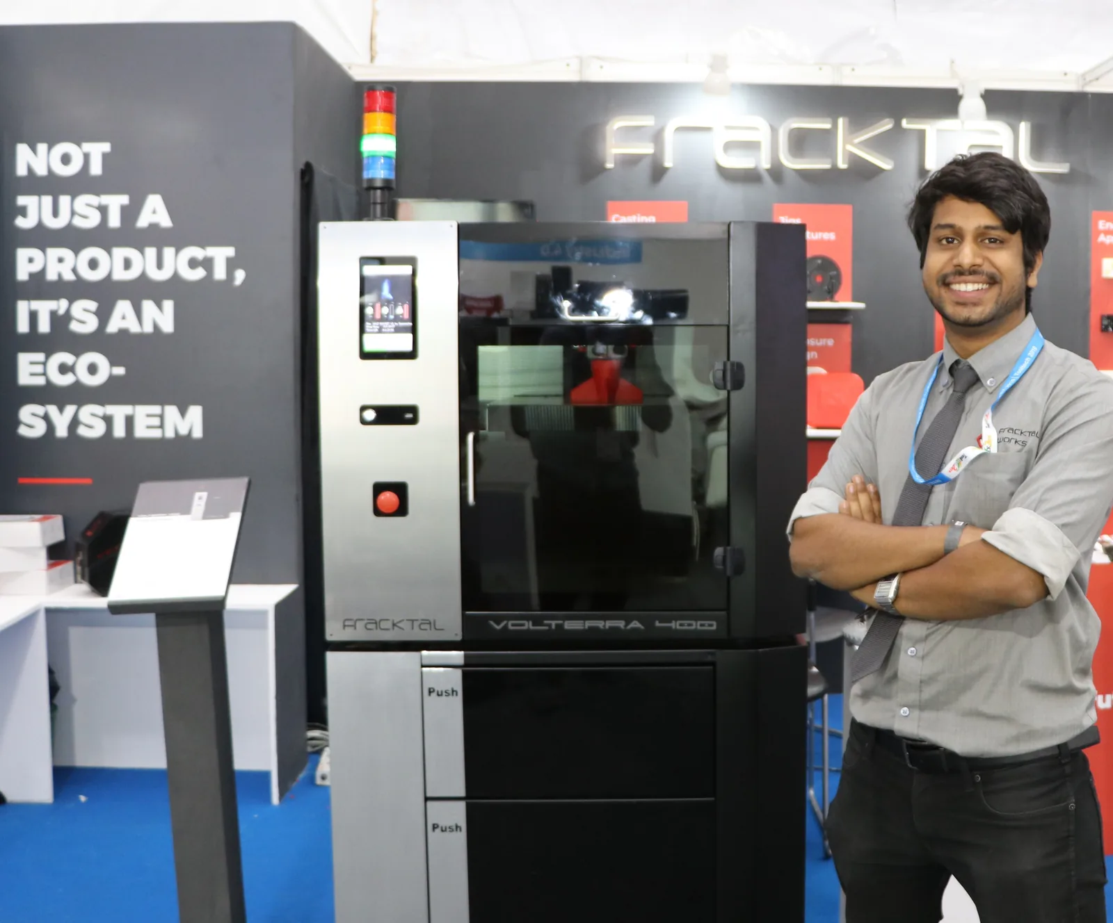 Vijay Raghav Varada, arms folded, beside a Fracktal Volterra 400 industrial 3D printer on the company's trade stand.
